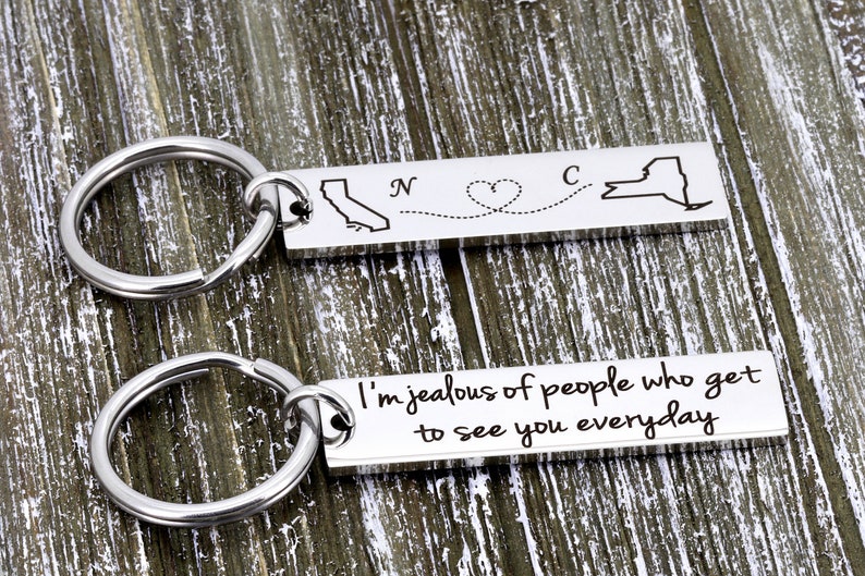 May include: Two silver keychains with a map of California and New York, connected by a dotted line with a heart. The second keychain has the text "I'm jealous of people who get to see you everyday".