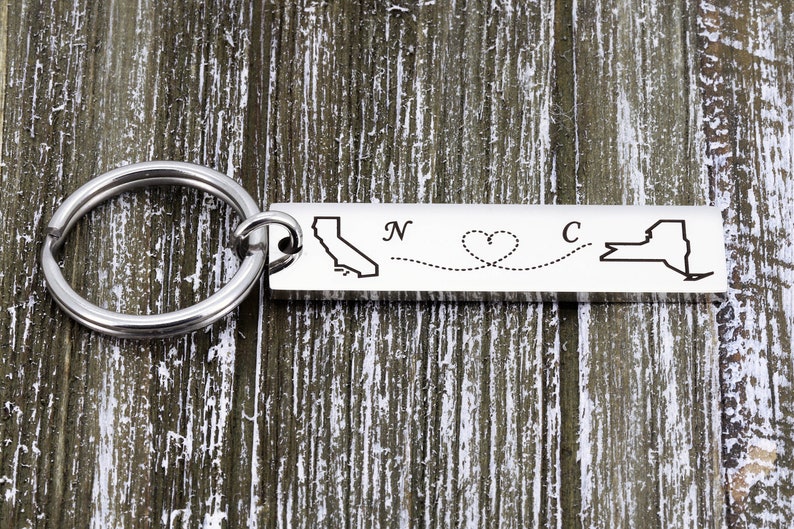 May include: Silver keychain with a map of California and New York connected by a dotted line with a heart in the middle. The text "N" and "C" are engraved on the keychain.