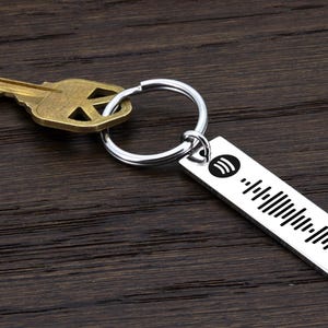 May include: A silver keychain with a Spotify logo and a barcode. The keychain is attached to a silver key ring and a house key.