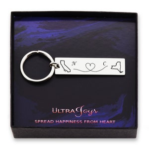 May include: Silver keychain with a map of California and New York connected by a dotted line with a heart in the middle. The text "N" and "C" are above the states. The keychain is in a black box with the text "ULTRA Joys" and "SPREAD HAPPINESS FROM HEART".