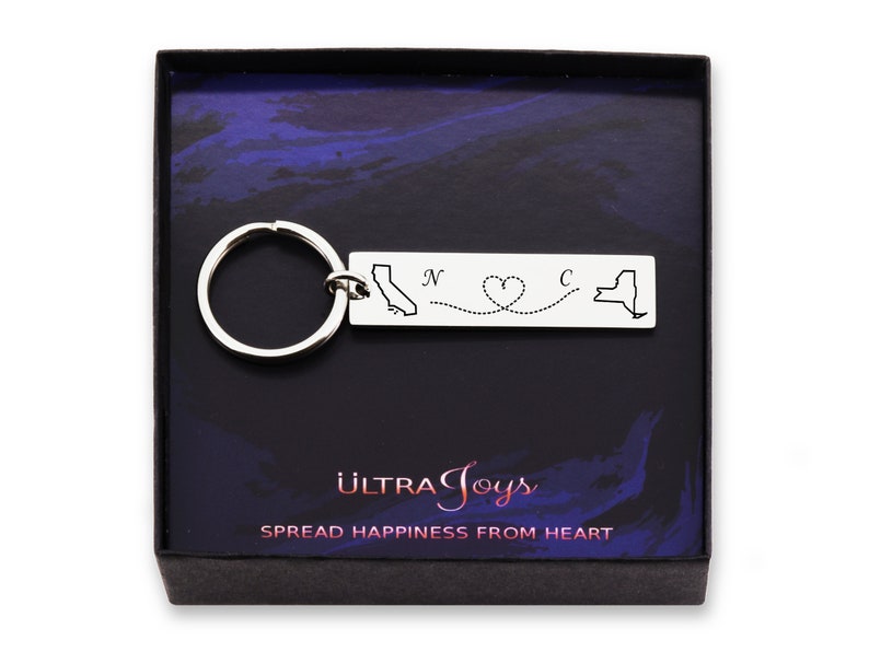May include: Silver keychain with a map of California and New York connected by a dotted line with a heart in the middle. The text "N" and "C" are above the states. The keychain is in a black box with the text "ULTRA Joys" and "SPREAD HAPPINESS FROM HEART".