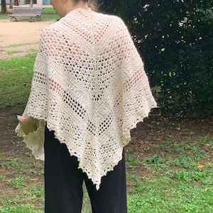 Puede incluir: Un chal blanco de ganchillo con un patrón de encaje. El chal está drapeado sobre los hombros de una persona y se lleva con pantalones negros.