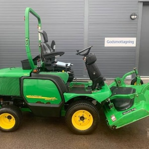 Puede incluir: Un cortacésped de conducción John Deere verde con una barra de seguridad, un asiento y una gran plataforma de corte. La cortadora está estacionada frente a un edificio gris con el texto "Godsmottagning" en el lateral.