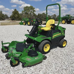 May include: A green John Deere riding lawn mower with a yellow seat and wheels. The mower has a black steering column and a cutting deck attached. Other green tractors are visible in the background under a blue sky.