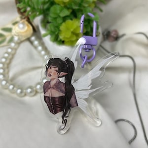 May include: A clear acrylic charm featuring a dark-haired fairy with white wings. The fairy is wearing a black corset and has a delicate, ethereal look. The charm is attached to a silver chain and a purple metal clasp.