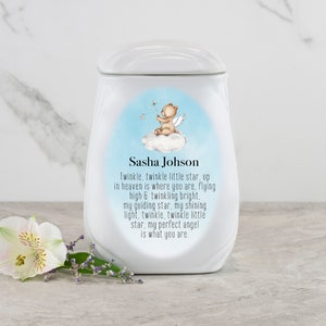 May include: White ceramic urn with a blue and white design featuring a teddy bear angel. The urn has the text "Sasha Johson" and a poem about a guiding star.
