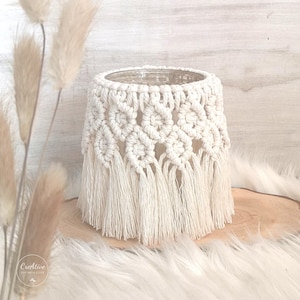 以下が含まれることがあります： オフホワイトのマクラメバスケット。円筒形で、下部にフリンジが付いています。バスケットは織り模様で、木製の表面に置かれています。全体的な美学はボヘミアンで手作りです。