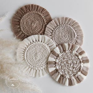 May include: Four macrame coasters in shades of beige and brown. Each coaster has a spiral center and a fringed edge. The coasters are arranged on a white surface, showcasing their handmade texture and design.