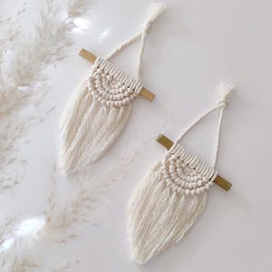 以下が含まれることがあります： オフホワイトのマクラメ壁掛け2つ。それぞれ、天然木のダボに取り付けられた織りの半円形デザインが特徴です。織り部分の下には、長く流れるフリンジが質感を加えています。吊り下げ用の結び目のあるコードが付いています。