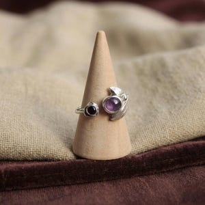 Può includere: Un anello d'argento con due pietre preziose, una nera e una viola, incastonate in un design a forma di foglia. L'anello è esposto su un piccolo supporto a forma di cono marrone chiaro, su uno sfondo di tessuto neutro. Il design dell'anello è dettagliato.