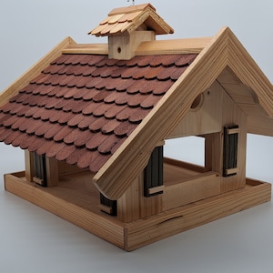 Puede incluir: Comedero para p&aacute;jaros de madera con un techo de tejas rojas y una peque&ntilde;a casa de madera en la parte superior. El comedero tiene una plataforma para que los p&aacute;jaros aterricen y una gran abertura para que los p&aacute;jaros accedan a la comida.