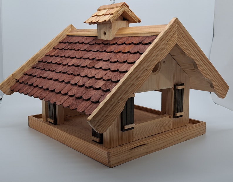 Puede incluir: Comedero para p&aacute;jaros de madera con un techo de tejas rojas y una peque&ntilde;a casa de madera en la parte superior. El comedero tiene una plataforma para que los p&aacute;jaros aterricen y una gran abertura para que los p&aacute;jaros accedan a la comida.