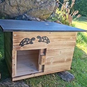 Può includere: Una casetta per tartarughe in legno rettangolare con un tetto piatto scuro e testurizzato e una struttura in legno chiaro. Il pannello frontale presenta due sagome decorative di tartarughe nere e un ingresso quadrato a sinistra.