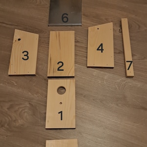 Könnte beinhalten: Sieben Holzstücke mit den Zahlen 1 bis 7 darauf gedruckt. Die Stücke sind in einem Muster auf einem Holzboden angeordnet.