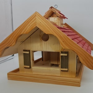 May include: Wooden bird feeder shaped like a house with a red shingled roof and a small, peaked roof on top. The feeder has two small windows with shutters and a landing platform below.