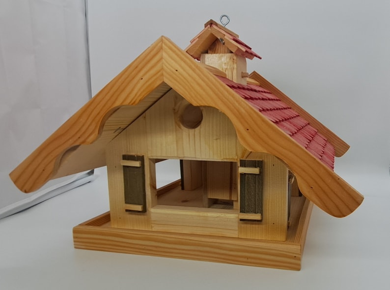 May include: Wooden bird feeder shaped like a house with a red shingled roof and a small, peaked roof on top. The feeder has two small windows with shutters and a landing platform below.