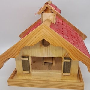 May include: A wooden bird feeder shaped like a house with a red shingled roof. The feeder has a small opening in the front and a perch on the bottom.