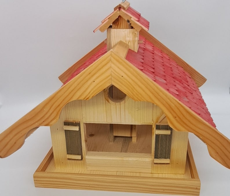 May include: A wooden bird feeder shaped like a house with a red shingled roof. The feeder has a small opening in the front and a perch on the bottom.