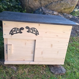 Può includere: Una casa di legno per tartarughe con tetto nero e porta scorrevole. Due sagome di tartarughe in metallo nero decorano la parte anteriore. La casa è realizzata in legno chiaro e poggia su una superficie erbosa.