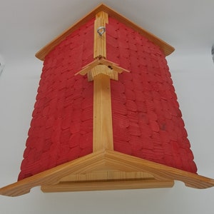 May include: A red wooden birdhouse with a shingled roof and a metal hook for hanging. The birdhouse has a simple design with a triangular roof and a rectangular base.