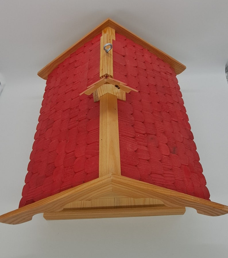 May include: A red wooden birdhouse with a shingled roof and a metal hook for hanging. The birdhouse has a simple design with a triangular roof and a rectangular base.