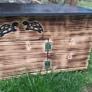 Op de afbeelding: Een rechthoekige houten kist met een geschroeide afwerking, voorzien van twee zwarte schildpadsilhouetten op het bovenste voorpaneel. De kist heeft een platte zwarte bovenkant en een scharnierende voordeur met twee zilveren metalen scharnieren.
