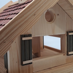 Puede incluir: Comedero para p&aacute;jaros de madera con un techo de tejas rojas, una peque&ntilde;a abertura redonda y una puerta con bisagras. El comedero tiene una base marr&oacute;n y un marco de madera clara.