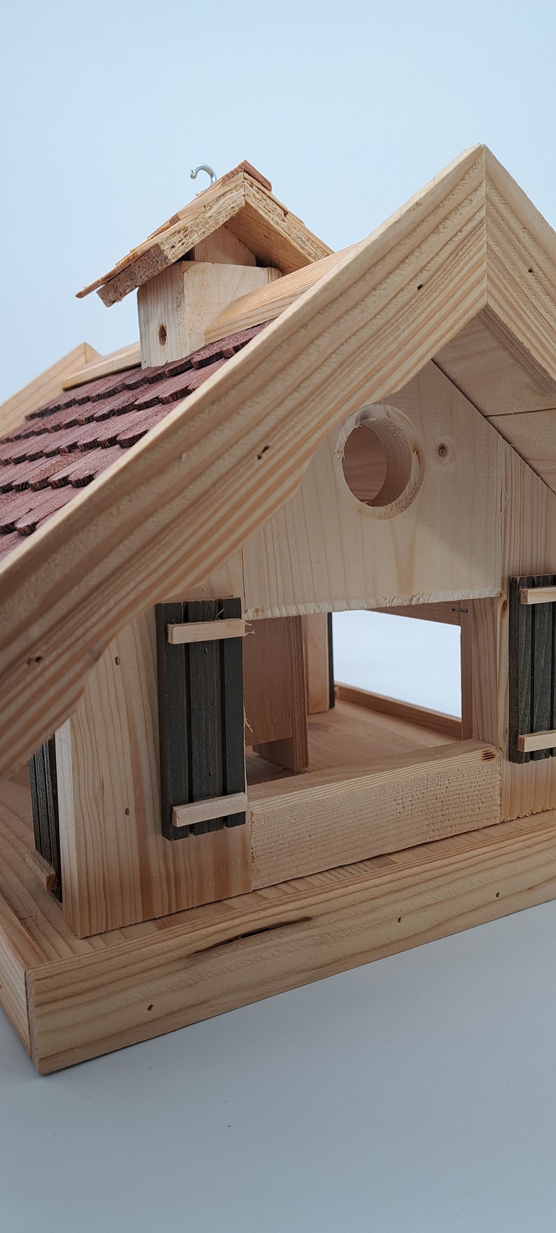 Puede incluir: Comedero para p&aacute;jaros de madera con un techo de tejas rojas, una peque&ntilde;a abertura redonda y una puerta con bisagras. El comedero tiene una base marr&oacute;n y un marco de madera clara.