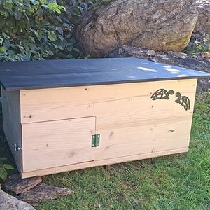 Könnte beinhalten: Ein hölzernes Schildkrötenhaus mit schwarzem Dach, einer kleinen Tür und zwei dekorativen Schildkrötensilhouetten. Das Haus besteht aus hellem Holz und steht auf einer Rasenfläche.