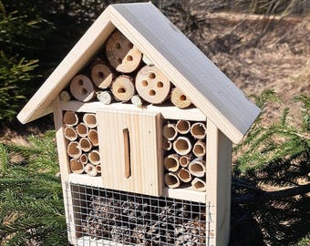 Puede incluir: Una casa de madera para insectos con múltiples compartimentos para diferentes tipos de insectos. La casa tiene una pequeña puerta y una sección inferior de malla. La casa está hecha de madera natural y tiene un color marrón claro.