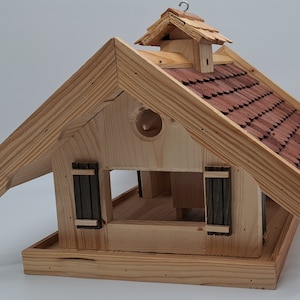 Puede incluir: Comedero para p&aacute;jaros de madera con forma de casita peque&ntilde;a con un tejado de tejas rojas. El comedero tiene una peque&ntilde;a abertura en la parte delantera y una percha en el lateral.