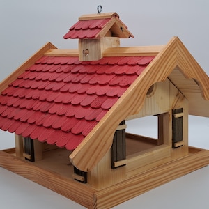 May include: A wooden bird feeder shaped like a small house with a red shingled roof. The feeder has a large opening in the front for birds to access the food.