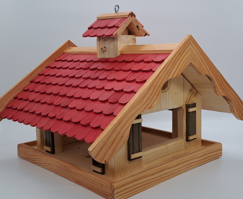 May include: A wooden bird feeder shaped like a small house with a red shingled roof. The feeder has a large opening in the front for birds to access the food.