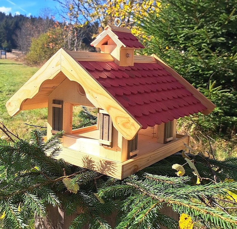 May include: A wooden bird feeder shaped like a small house with a red roof. The feeder has two small windows and a perch on the front.
