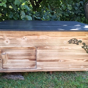 Op de afbeelding: Een houten schildpaddenverblijf met een donker dak en een kleine deur. Het huis is gemaakt van lichtbruin hout met een decoratief schildpadontwerp. Het huis staat op groen gras.