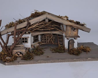 Puede incluir: Un modelo de madera de una pequeña casa rústica con techo de paja y exterior encalado. La casa tiene una pequeña puerta y ventanas, y hay un árbol delante de ella. El modelo está colocado sobre una base de madera.
