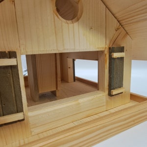 May include: A wooden bird feeder with a pitched roof and two small green shutters. The feeder has a platform for birds to land on and a small opening for birds to access the food.