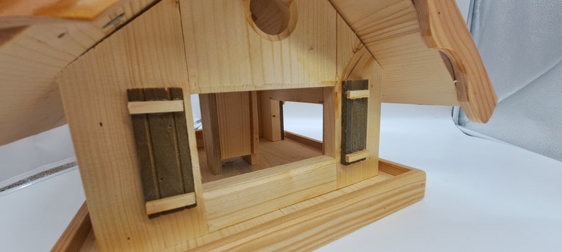 May include: A wooden bird feeder with a pitched roof and two small green shutters. The feeder has a platform for birds to land on and a small opening for birds to access the food.