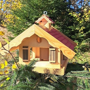 May include: A wooden bird feeder shaped like a small house with a red roof. The feeder has two small windows with shutters and a perch on the front.