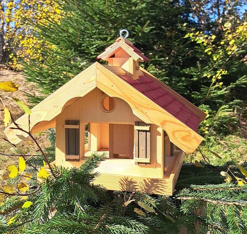 May include: A wooden bird feeder shaped like a small house with a red roof. The feeder has two small windows with shutters and a perch on the front.