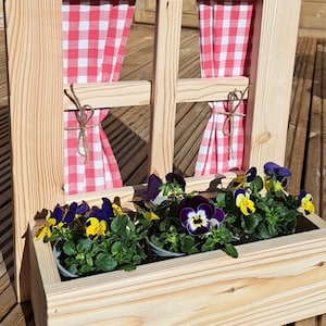 Peut inclure: Une jardinière en bois en forme de fenêtre avec des rideaux à carreaux rouges et blancs. La jardinière est remplie de plantes vertes et de pensées violettes et jaunes.