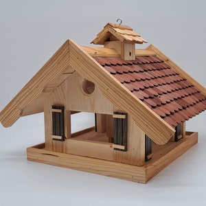 Puede incluir: Comedero para p&aacute;jaros de madera con forma de casa con un techo de tejas rojas. El comedero tiene dos ventanas peque&ntilde;as y una percha en la parte delantera.