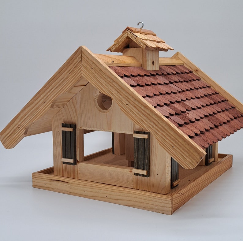 Puede incluir: Comedero para p&aacute;jaros de madera con forma de casa con un techo de tejas rojas. El comedero tiene dos ventanas peque&ntilde;as y una percha en la parte delantera.