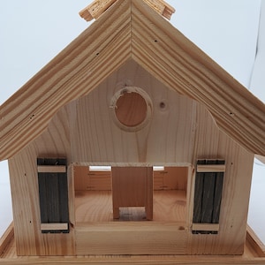 Puede incluir: Casa de p&aacute;jaros de madera con techo a dos aguas y una abertura circular. La casa de p&aacute;jaros tiene dos peque&ntilde;as puertas en la parte delantera y una plataforma debajo.