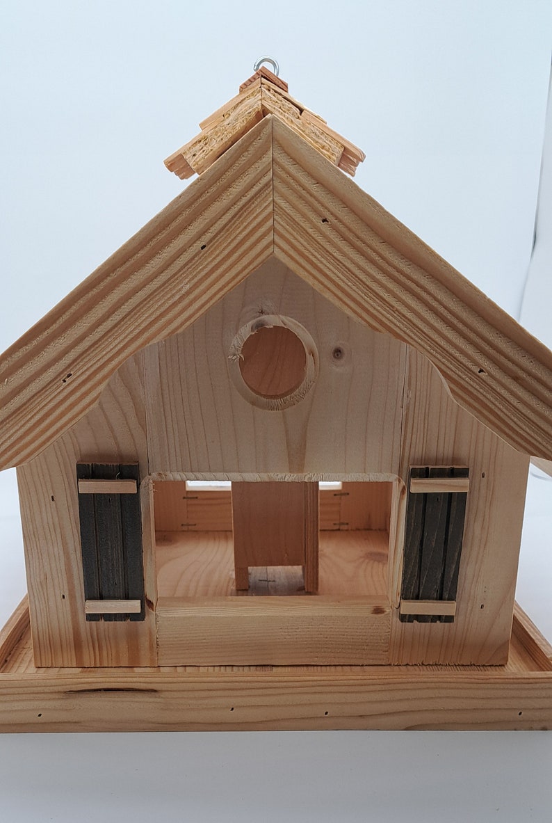 Puede incluir: Casa de p&aacute;jaros de madera con techo a dos aguas y una abertura circular. La casa de p&aacute;jaros tiene dos peque&ntilde;as puertas en la parte delantera y una plataforma debajo.