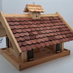 Puede incluir: Un comedero para p&aacute;jaros de madera con forma de casita con un tejado de tejas rojas. El comedero tiene una peque&ntilde;a plataforma debajo para que los p&aacute;jaros puedan posarse.