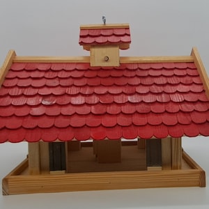 May include: A wooden bird feeder with a red shingled roof and a small, peaked roof on top. The feeder has a platform for birds to land on and a tray for food.