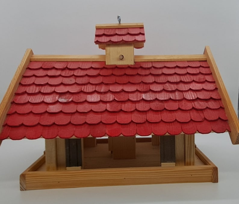 May include: A wooden bird feeder with a red shingled roof and a small, peaked roof on top. The feeder has a platform for birds to land on and a tray for food.