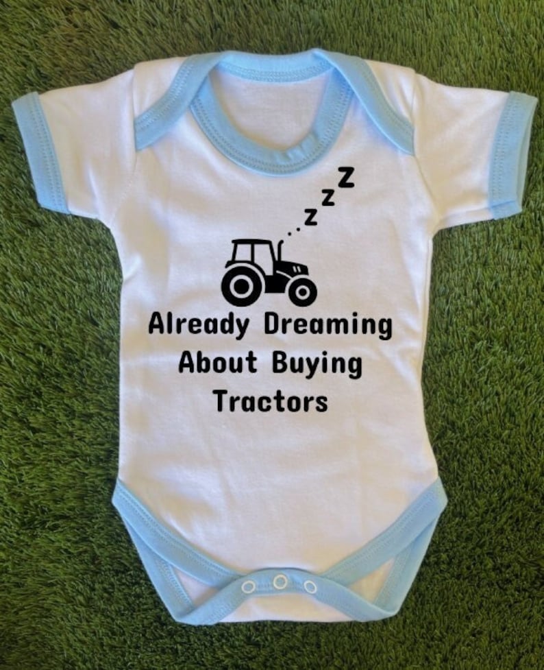 May include: White baby onesie with blue trim. The onesie has a black tractor graphic and the text "Already Dreaming About Buying Tractors" with three Z's above the tractor.