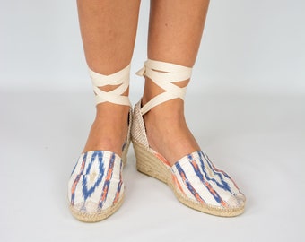 May include: Espadrille wedge sandals with a cream-colored fabric upper featuring a blue and orange ikat pattern. The sandals have a woven jute sole and cream-colored ankle ties.